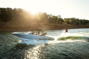 A person wakesurfs behind a 2026 boat on a lake, sunlight filtering through trees on the shoreline in the background—an exciting scene you might find at the Supreme Boat Show or during a major Boat Sales Event.