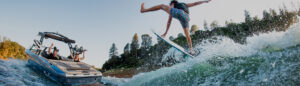 A person performs a trick on a wakesurf board behind a Supreme Wake Boat on a lake, with trees and blue sky in the background.