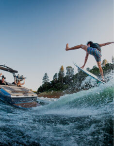 A person on a surfboard performs a jump on a lake while people on a nearby Supreme Wake Boat watch and take photos.