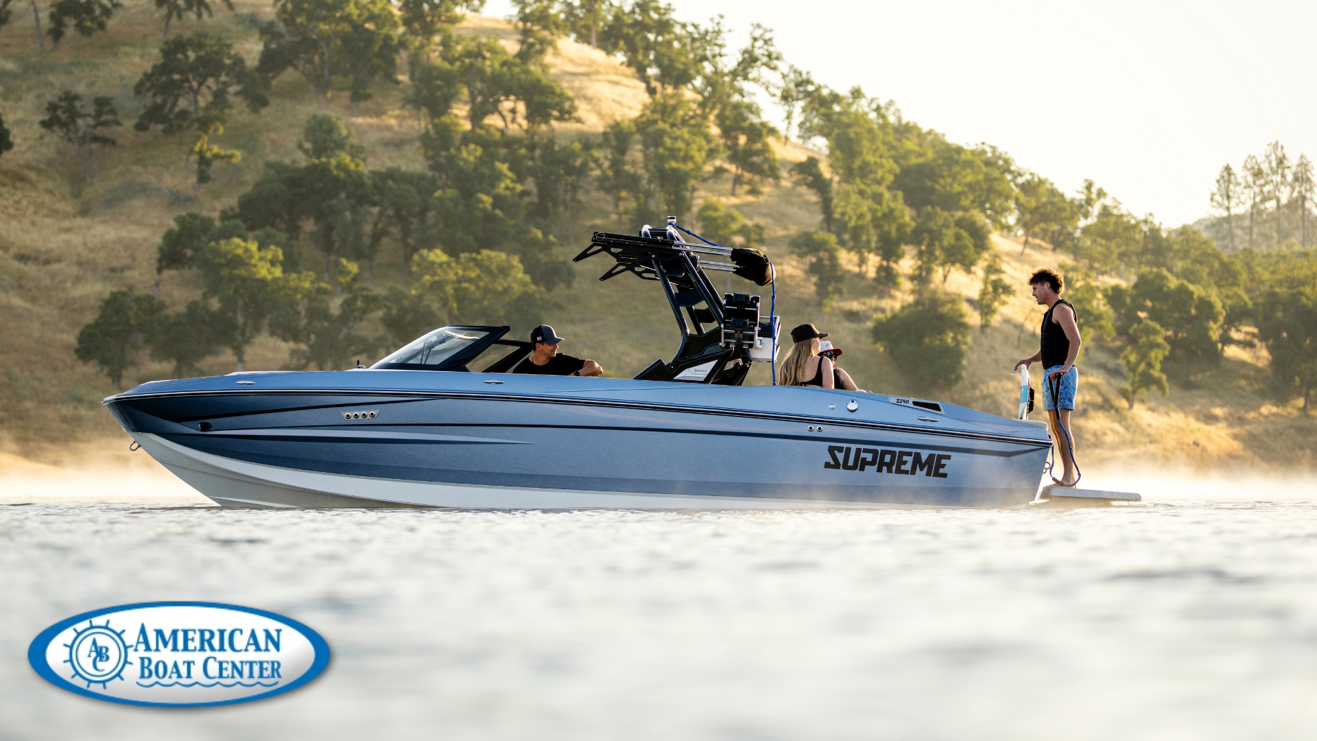 A blue Supreme Boats speedboat with several people on board floats on a lake near tree-covered hills; American Boat Center logo is visible in the corner.