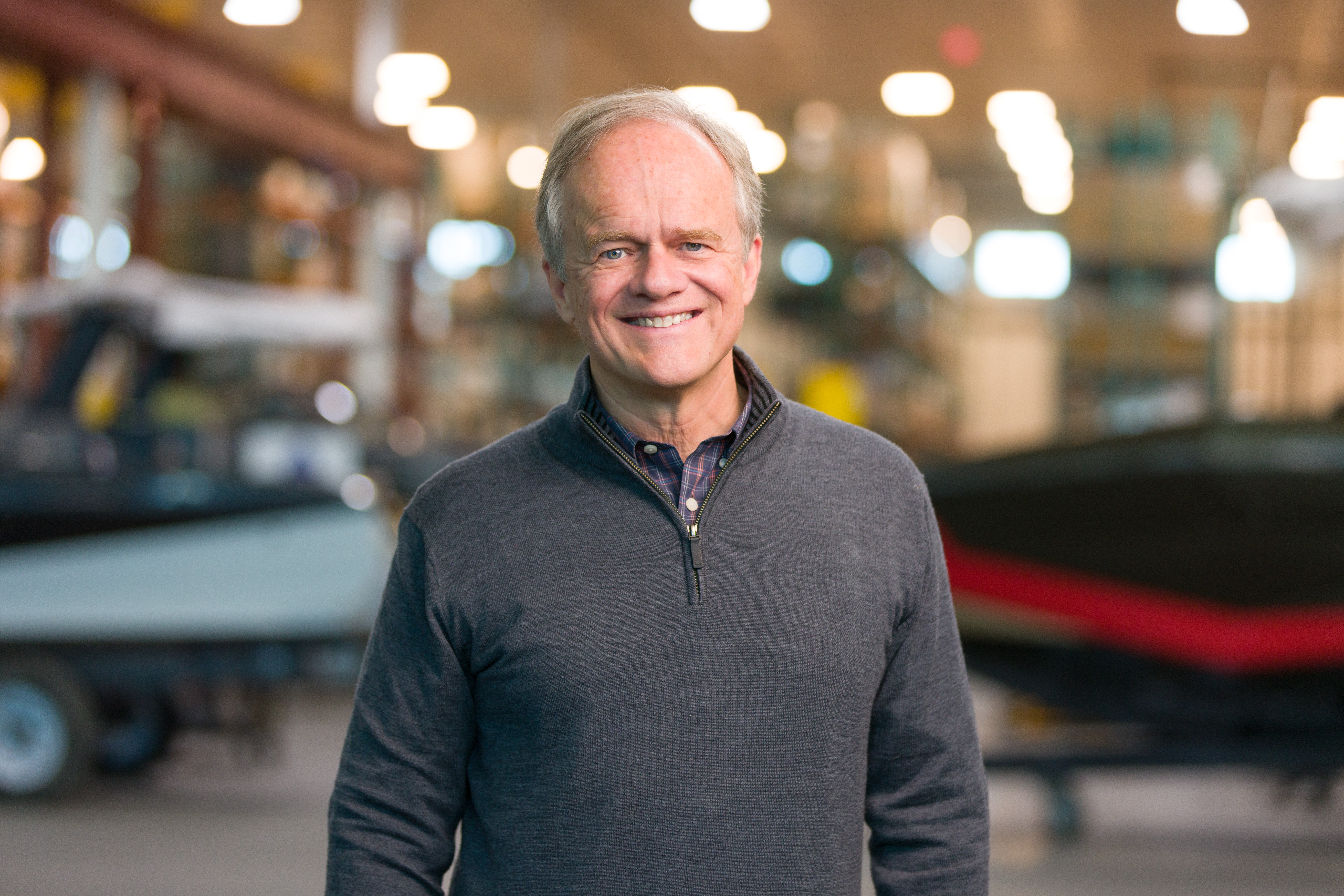 Older man with gray hair wearing a gray zip-up sweater stands indoors in a workshop or warehouse, with blurred shelves and Supreme Boats in the background.