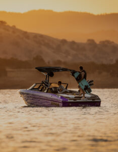 A purple Supreme Wake Boat with two people on board cruises a lake at sunset; one person holds a wakeboard while hills rise in the background.