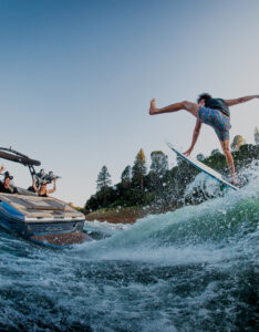 A person wakesurfs on a lake near a Supreme Wake Boat filled with cheering passengers, with trees and a clear sky in the background.