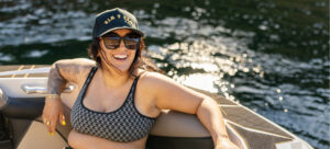 Woman wearing sunglasses and a black cap sits smiling on a boat, with water and sunlight in the background—an effortless snapshot of The Supreme Life.