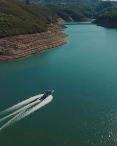 A motorboat moves across a wide greenish-blue river, leaving a wake behind, as you Experience Supreme Boats along hilly, forested banks on either side under clear skies.