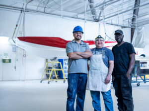 Three workers stand side by side in a bright industrial workshop, with a partially painted red and white boat suspended in the background, capturing a moment from an engaging factory tour.