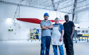 Three people wearing work clothes and safety gear stand smiling in a spacious industrial workshop, as if ready to welcome guests on a factory tour, with boats and equipment visible in the background.