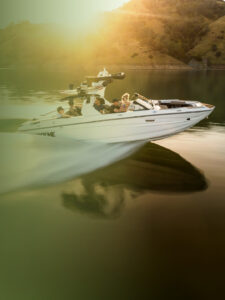 A white Supreme wakeboat with several people onboard moves quickly across a calm lake, with sunlight and hills in the background.
